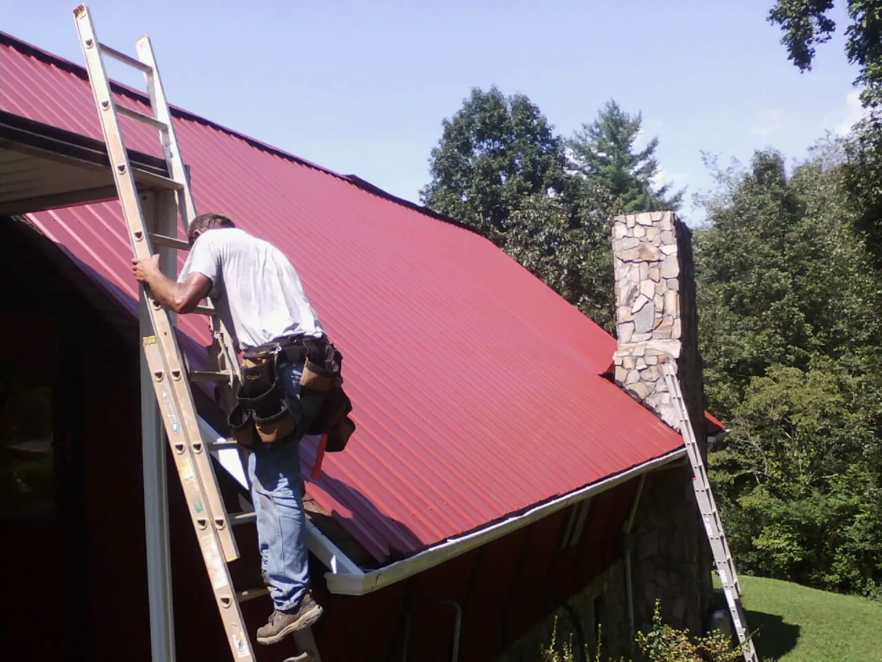 Roofer on a ladder beside a red metal roof and stone chimney — Soffit & Fascia Repair by Roofing Brothers in Guatay